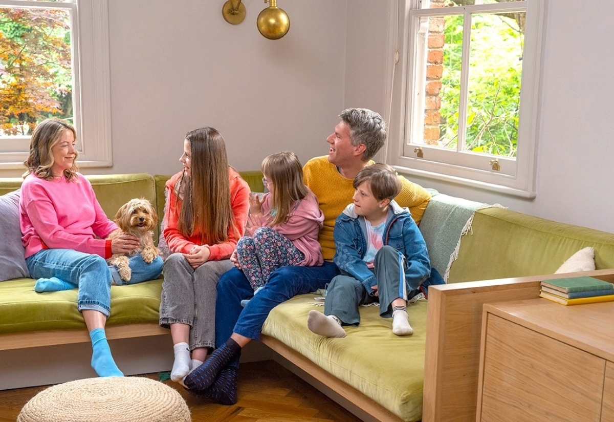 A family and their dog enjoy time together seated on a couch in front of a large window.