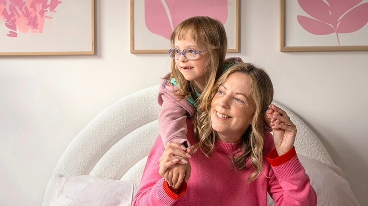 A young child playfully drapes their arms over an adult's shoulders as they sit together on a bed.