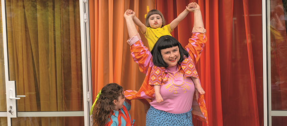 A woman joyfully holds up her arms with a smiling child on her shoulders.
