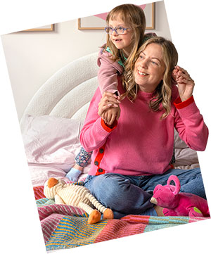 A young child playfully drapes their arms over an adult's shoulders as they sit together on a bed.