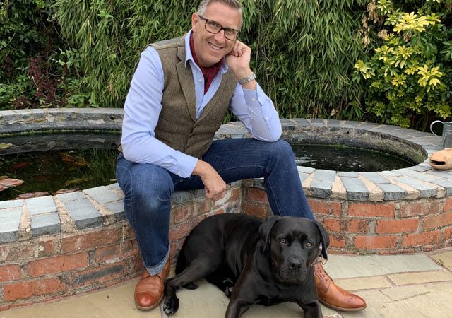 Graeme Hall, television presenter and author, sitting by a garden pond with a black labrador resting at his feet.