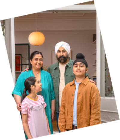 Two adults and two children stand close together outside a home, all smiling and dressed casually.