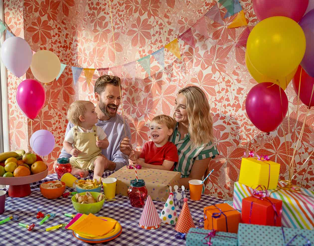 Two adults and two children, sitting at a decorated table with balloons, wrapped gifts and party items around them.