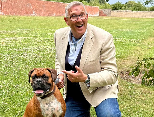 Graeme Hall, known as The Dogfather, kneeling on grass in a walled garden, smiling and pointing toward a Boxer dog.