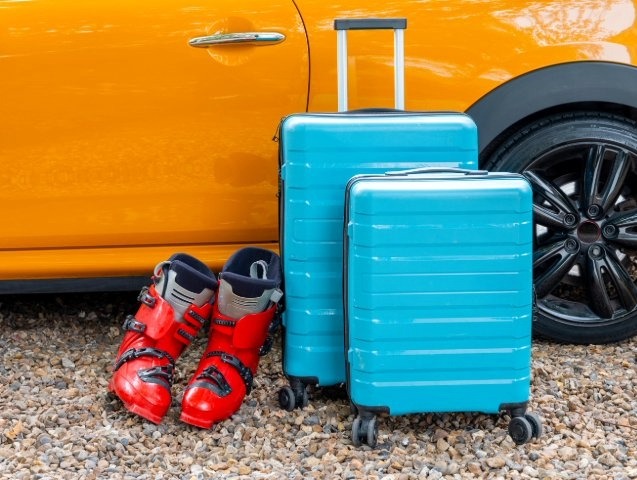 A suitcase and ski boots are placed next to a parked car, ready for a winter adventure.